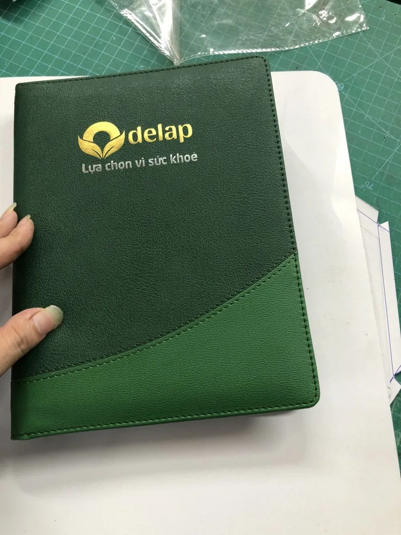 Sổ b&igrave;a xanh c&oacute; logo kh&aacute;ch h&agrave;ng được cầm cạnh tập giấy tr&ecirc;n b&agrave;n l&agrave;m việc
