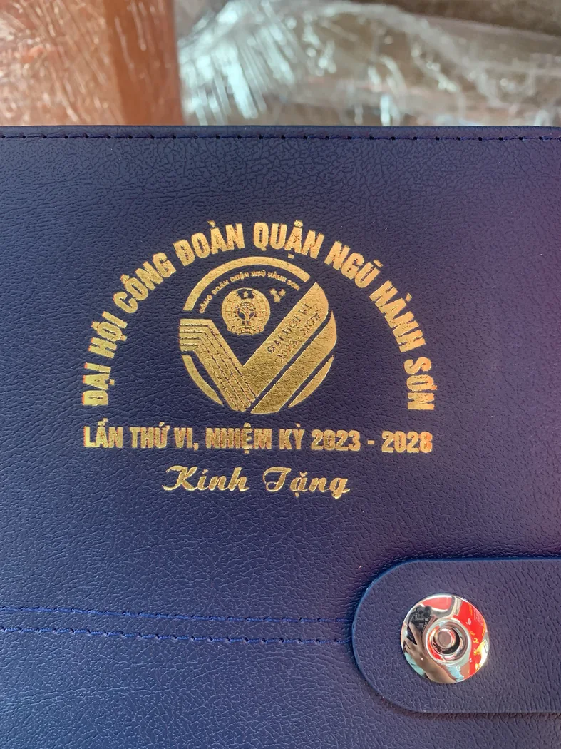 Logo v&agrave; d&ograve;ng chữ m&agrave;u v&agrave;ng in tr&ecirc;n b&igrave;a sổ xanh đậm c&oacute; v&acirc;n v&agrave; đường may