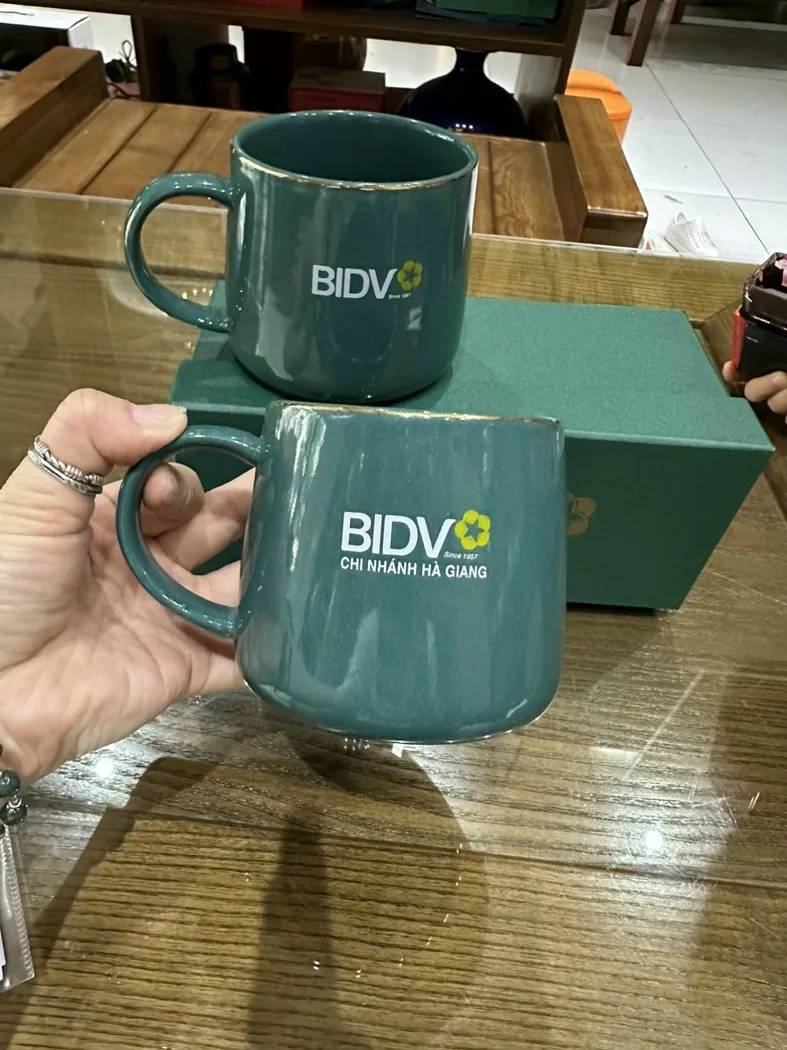 Hai cốc sứ m&agrave;u xanh in logo BIDV v&agrave; t&ecirc;n chi nh&aacute;nh kh&aacute;c nhau