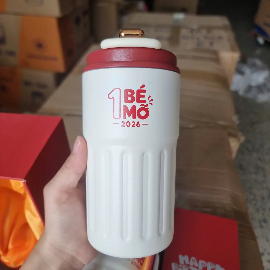 Cốc giữ nhiệt trắng in chữ chương tr&igrave;nh qu&agrave; cầm tr&ecirc;n tay cạnh hộp đỏ