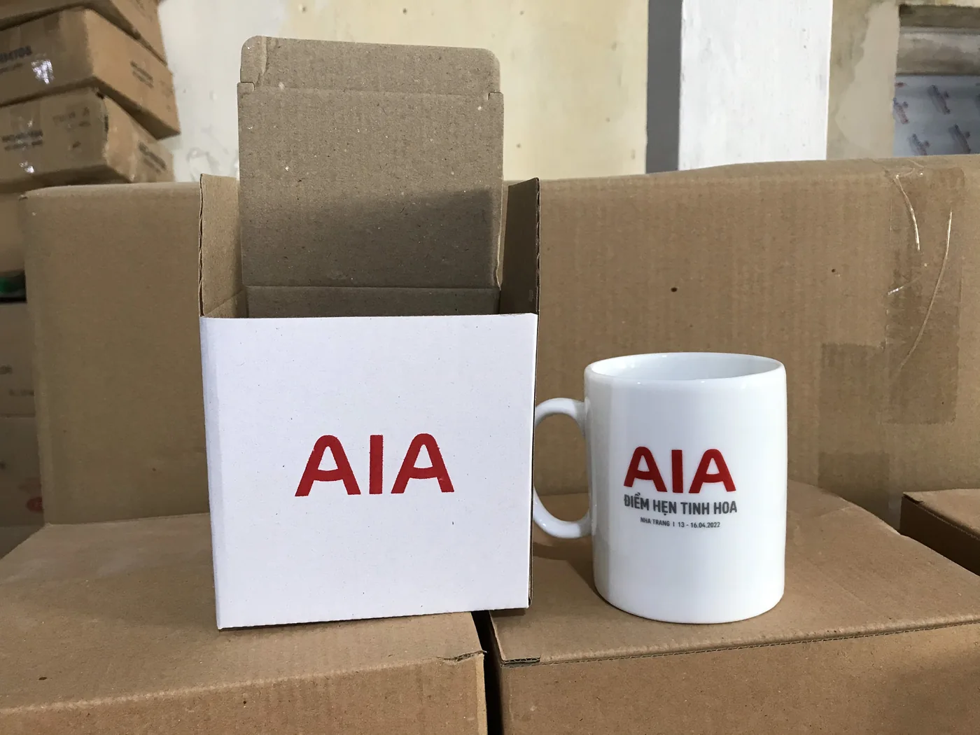 Cốc sứ v&agrave; hộp giấy AIA đặt tr&ecirc;n th&ugrave;ng carton