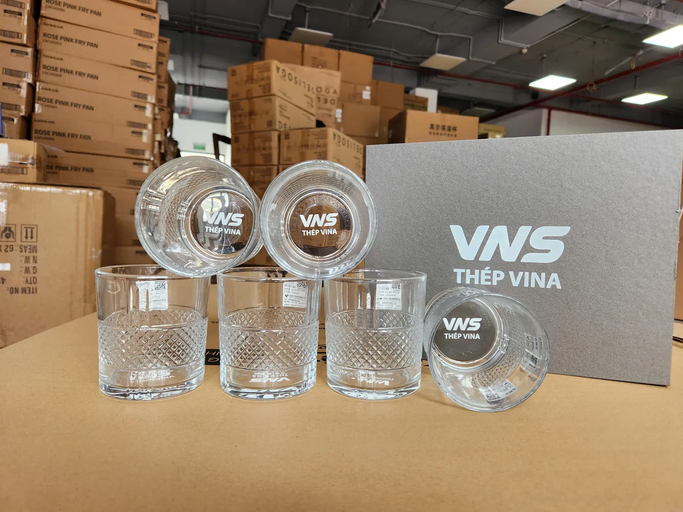 C&aacute;c cốc thủy tinh in logo VNS đặt trước hộp x&aacute;m c&ugrave;ng nh&atilde;n, cạnh tem d&aacute;n tr&ecirc;n một số cốc