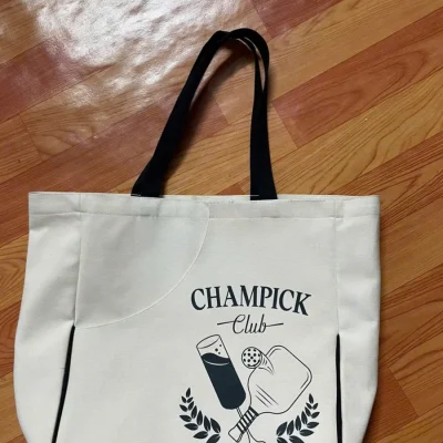 Ảnh thật túi canvas quai đen có hình CHAMPICK Club trên nền gỗ